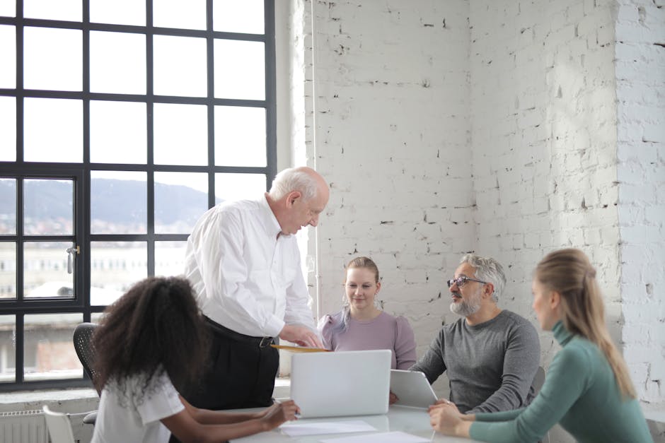 Concentrated adult man explaining business report to colleagues during brainstorm in contemporary office with laptop and tablet near window looking at each other